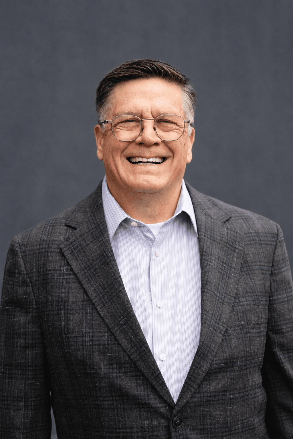 Mark Norman, laughing warmly, in a gray plaid blazer photographed against a charcoal backdrop.