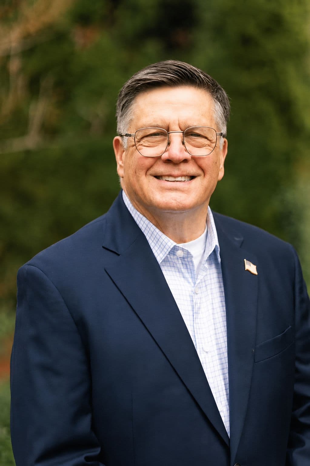 Mark Norman, smiling, in a navy blazer with an American flag lapel pin, photographed outdoors against a green backdrop.