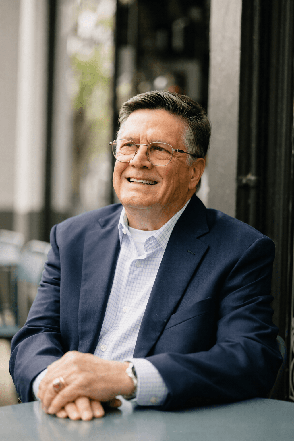 Mark Norman, seated outside a café in a navy blazer, smiling warmly with hands clasped on the table.