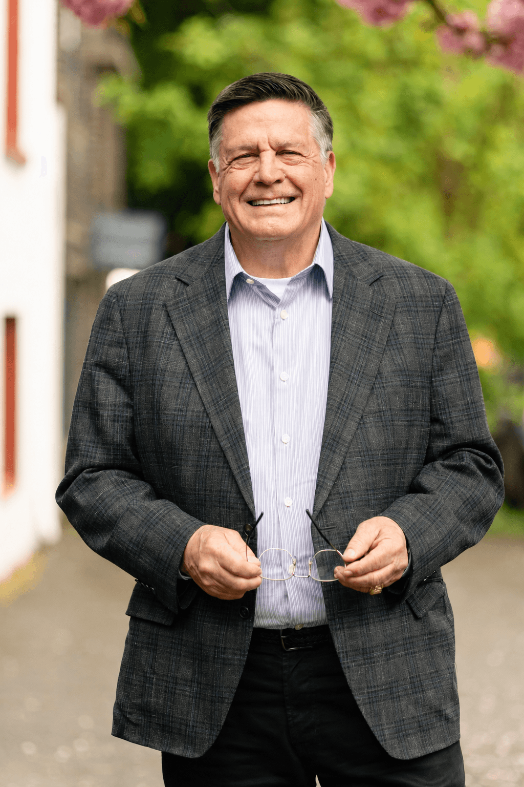 Mark Norman, smiling, holding his glasses, in a gray plaid blazer photographed outdoors with cherry blossoms in the background.