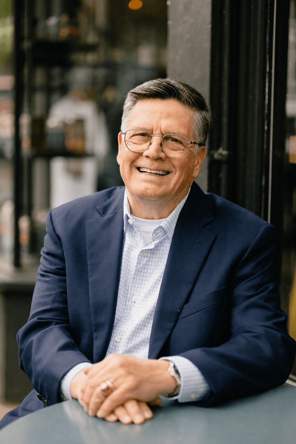 Mark Norman, seated outside a café, laughing warmly with hands clasped, in a navy blazer.